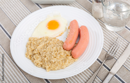 Obraz na płótnie Two sausages with a fried egg and oatmeal on a white plate. Breakfast option with eggs and porridge next to cutlery and a glass of water w salonie