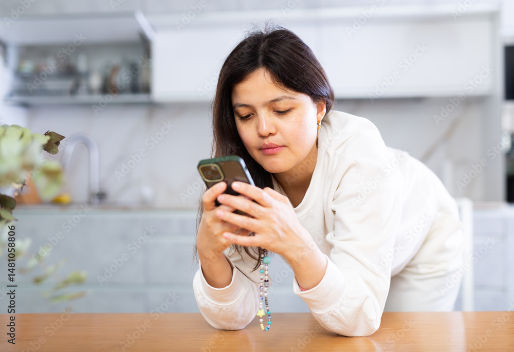 Relaxed young dark-haired woman dressed in casual white clothing standing leaning on table in home kitchen, scrolling through social media on smartphone