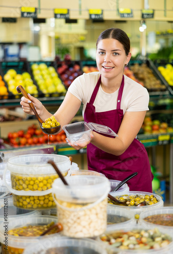 Young woman seller in apron lays out olives in supermarket
