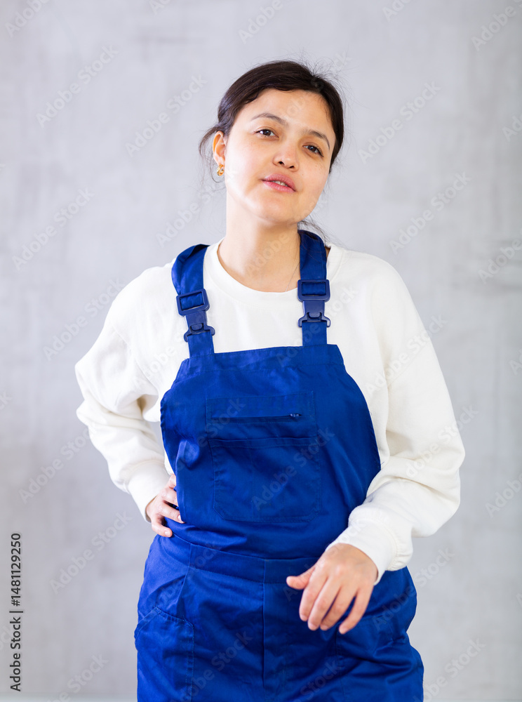 Portrait of young female mechanic in blue overalls posing in studio against gray plain background, looking confident and positive. Professional activity concept