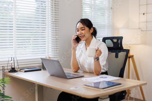 Happy young business woman manager talking consulting client on cell phone working on laptop in the office. Busy woman professional business making call on cell using computer sitting at desk.