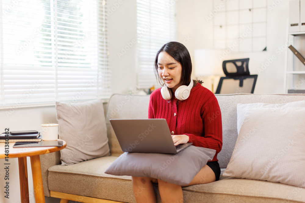 Happily asian woman wearing headphones holding working from home on her laptop while lying on sofa in bright apartment cheerful remote work lifestyle. Happily asian woman wearing headphones holding working from home on her laptop while lying on sofa in bright apartment cheerful remote work lifestyle.