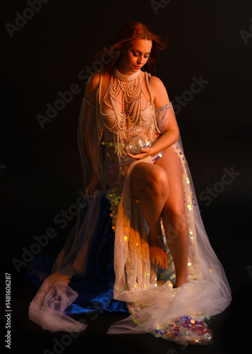Fantasy studio portrait of a red-haired mermaid faery model wearing a white pearl gown, fairytale costume. posing in a classical sitting position with flowing fabric and ethereal moody studio lighting
