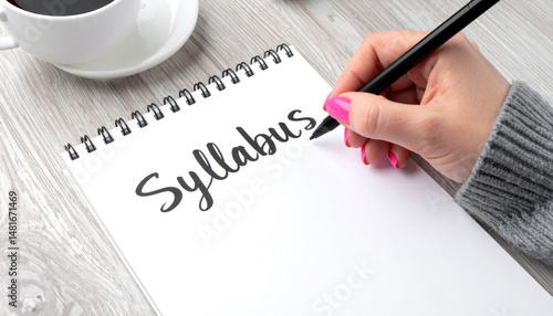 Woman hand writes Syllabus on white notepad, wearing grey sweater, with coffee cup nearby