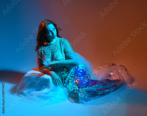 Fantasy studio portrait of a red-haired mermaid faery model wearing white pearl gown, fairytale costume. classical sitting pose, with flowing fabric, colourful neon moody lighting on studio background