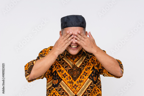 Smiling Indonesian man playfully covering his eyes with both hands, showing cheerful mood in Batik shirt and headwear. Isolated on a light background.