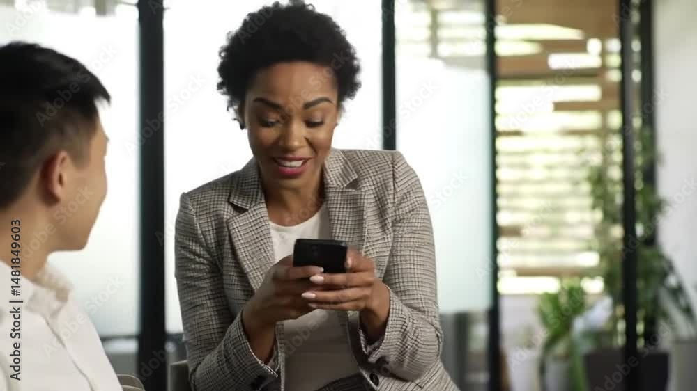 Happy African American businesswoman using mobile phone while talking to her colleague in waiting room Happy African American businesswoman using mobile phone while talking to her colleague in waiting room