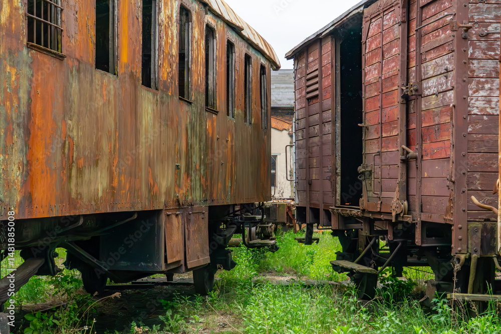 Obraz premium Old railway cars standing on a siding, covered with rusty metal and weathered wood. Visible signs of time and neglect create a strong atmosphere of decay and abandonment. Railway buildings can be seen