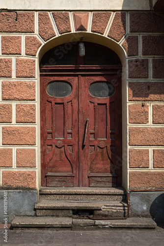Wallpaper Mural Worn red wooden double doors framed by a textured archway glow in warm sunlight. Cracks, scratches, and old stone steps whisper stories of age, mystery, and quiet European charm. Torontodigital.ca