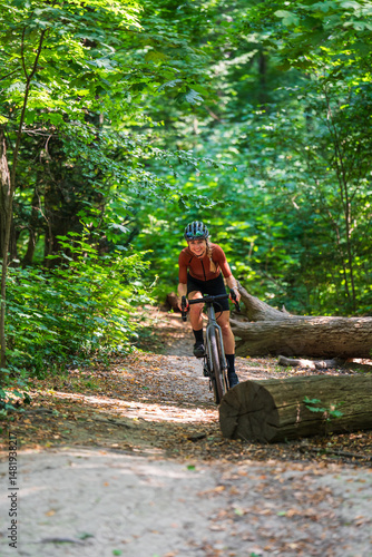 Happy cyclist in sports uniform, helmeted, rushing on gravel bike on forest path among fallen trees. Active recreation, healthy lifestyle
