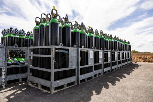 Wallpaper Mural Numerous green and white topped black gas cylinders are secured in metal crates and stored outdoors on a paved surface under a partly cloudy sky Torontodigital.ca