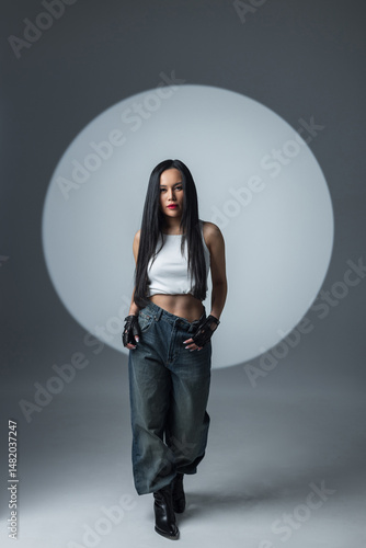Cool urban style – woman in crop top, jeans, and boots walks under a circular spotlight in a modern studio.