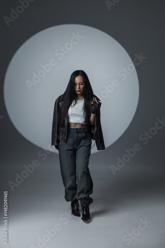 Confident woman in edgy streetwear poses in a dramatic spotlight. Wearing a leather jacket, loose jeans, and gloves, she exudes power and attitude.