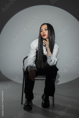 Beautiful fashion asian woman with red lips in trendy stylish outfit with white shirt and black corset sits on chair on dark background in photo studio. Pretty girl