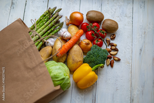 Fresh vegetables and fruits spilling from grocery bag. Top view of assorted fresh vegetables, fruits, and nuts spilling from a brown paper grocery bag onto a rustic wooden table.