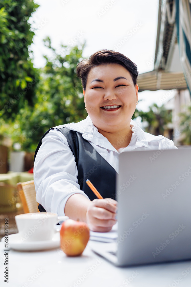 Fototapeta premium Smiling Asian woman working on a laptop at a cafe with a cup and apple during sunny weather