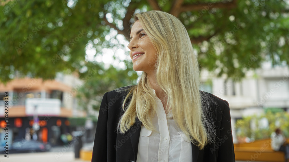 Fototapeta premium Woman smiling outdoors in urban park setting with green trees and city buildings, showcasing a confident blonde adult enjoying the vibrant street atmosphere.