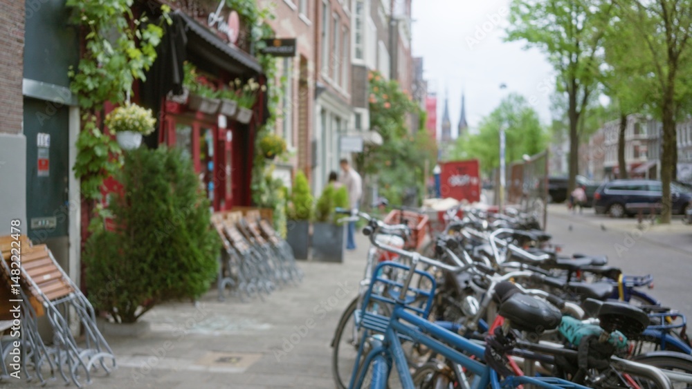 Obraz premium Blurred bicycles line a charming amsterdam street with vibrant shops and greenery, capturing the essence of urban life in the netherlands.