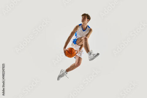 Φωτογραφία Young athletic man, basketball player soaring upward with dynamic posture, gripping ball in preparation for dunk against white background