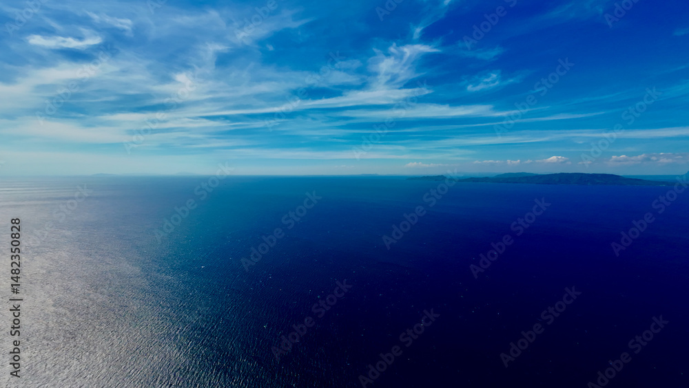 Obraz premium Aerial view of the sea surface and clouds above the horizon. Aerial view of the calm surface of the blue ocean, white clouds above the horizon and a small tropical island in the distance.