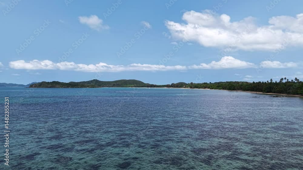 Drone captures wide coastal scene with gentle ocean waves moving toward tropical island shoreline under bright blue sky while distant green hills stretch along tranquil sea horizon. Drone captures wide coastal scene with gentle ocean waves moving toward tropical island shoreline under bright blue sky while distant green hills stretch along tranquil sea horizon.