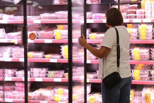 Fotografie Rear view of adult female customer opens big glass door in supermarket's fridge and chooses packaged fresh chilled meat