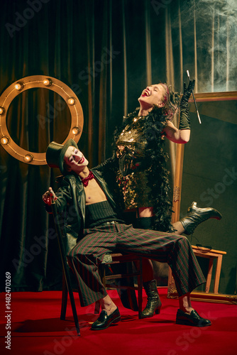 Expressive laughter and dramatic poses of two cabaret performers in vintage costumes on confetti-covered red carpet with smoke, lively moment. Concept of cabaret, show, circus, retro, vintage