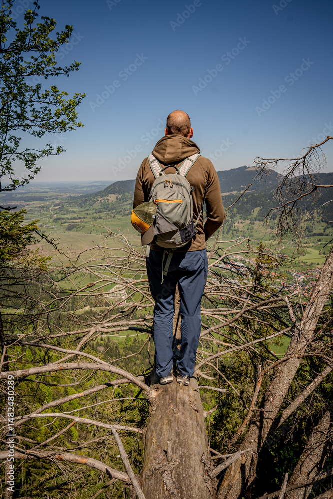 Naklejka premium Adventurous Hiker Exploring Naturpark Ammergauer Alpen: Lone Figure Standing On Rocky Outcrop Overlooking Majestic Mountain Ridges And Lush Green Valley Under A Clear Blue Sky In Scenic Bavaria