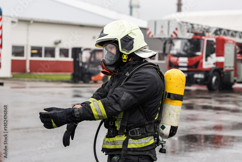 Wallpaper Mural Firefighter in full protective gear adjusts gloves during a rainy day at a fire station. Fire truck and buildings visible in the background Torontodigital.ca