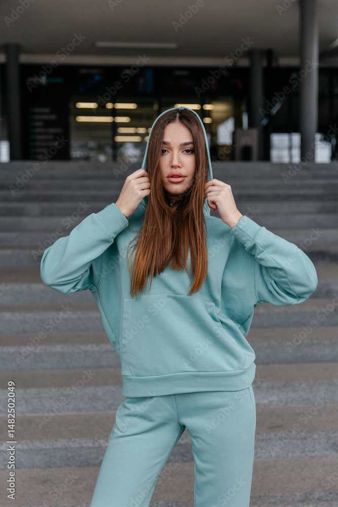 Fototapeta premium Cute girl model in a sporty comfortable casual turqouise outfit with a hoodie and sweatpants posing on the street