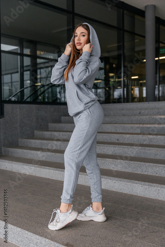 Cute girl model in a sporty comfortable casual gray outfit with a hoodie and sweatpants posing outdoor