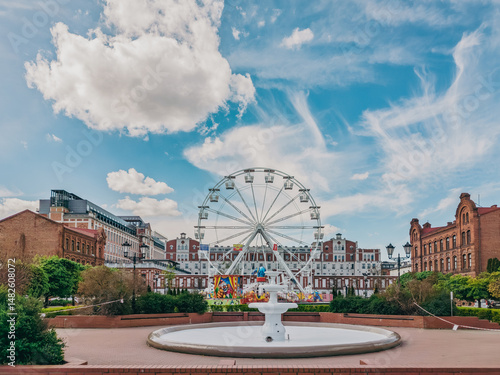 Ferris wheel and a non-operational fountain in a town square in Żyrardów, Poland, surrounded by brick buildings under a partly cloudy sky.