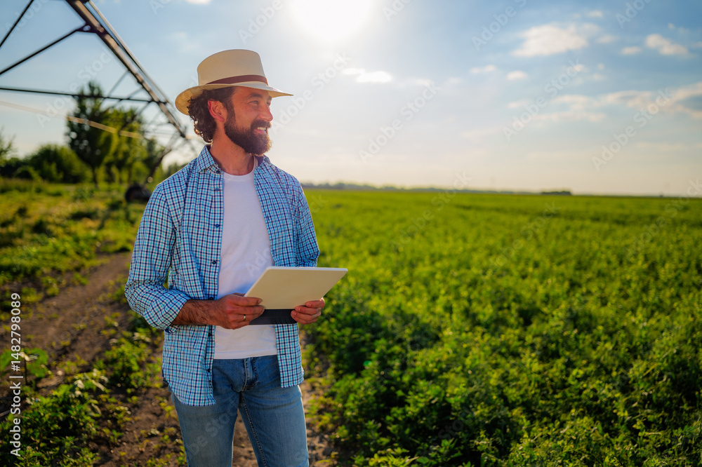 Fototapeta premium Confident farmer holding a digital tablet while admiring crops in a cultivated field with an irrigation system, highlighting modern technology for sustainable agriculture