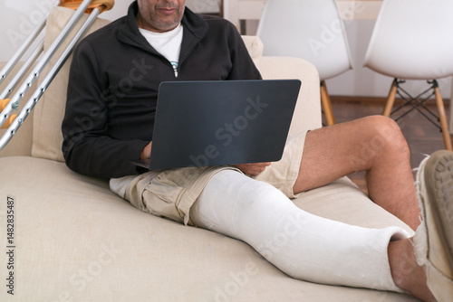 Man with broken leg using laptop on sofa with crutches nearby