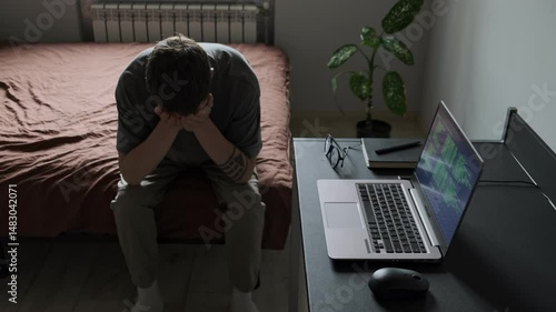 Depressed man with head down sitting on bed in despair in home office next to open laptop and glasses, illustrating mental fatigue. Realistic depiction of freelance burnout.
