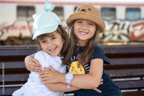 Two happy little sisters or best friends hugging and smiling at a bench. Adorable children enjoying, friendship concept.