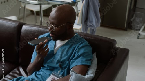 Young relaxed African American male doctor in eyeglasses and blue uniform sitting on sofa after work and talking to friend or family while using speakerphone