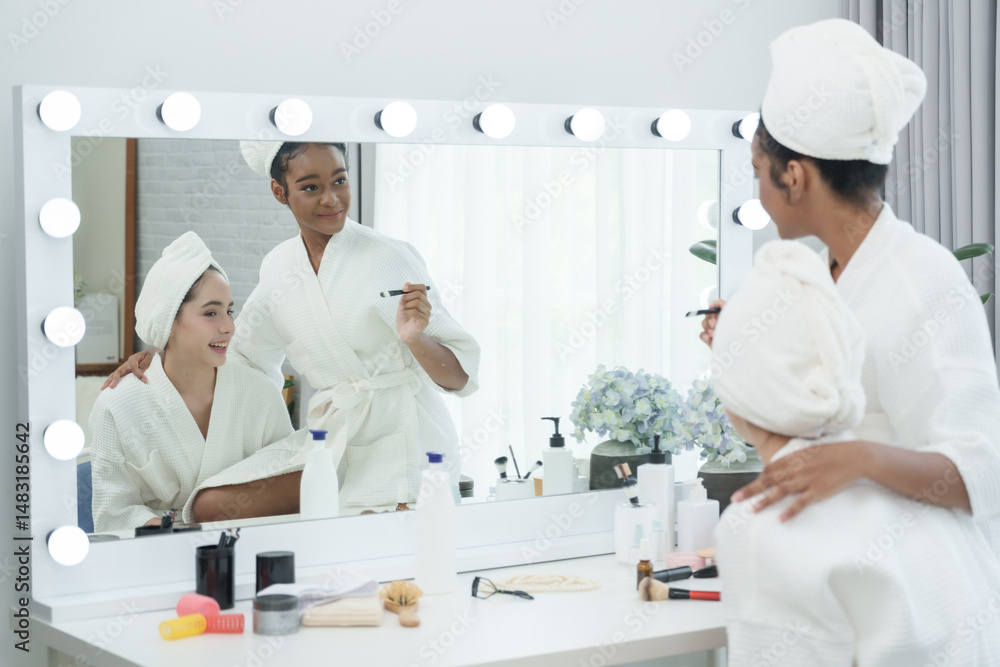 Fototapeta premium Two teenage girls in spa robes looking in mirror during makeup session, one African American and one mixed Caucasian Asian, expressing feminine diversity beauty routine and joyful lifestyle