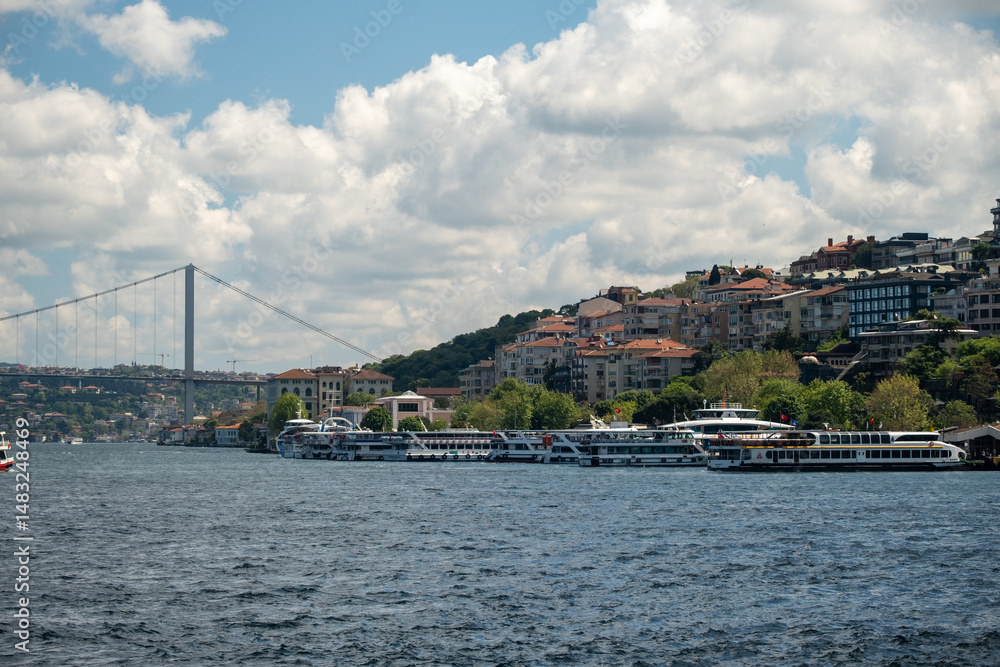 Naklejka premium Fishing boats on the Bosphorus with large ships passing under the Bosphorus Bridge in Istanbul, Turkey on a bright day with scenic clouds, hills, and a Turkish flag in the background