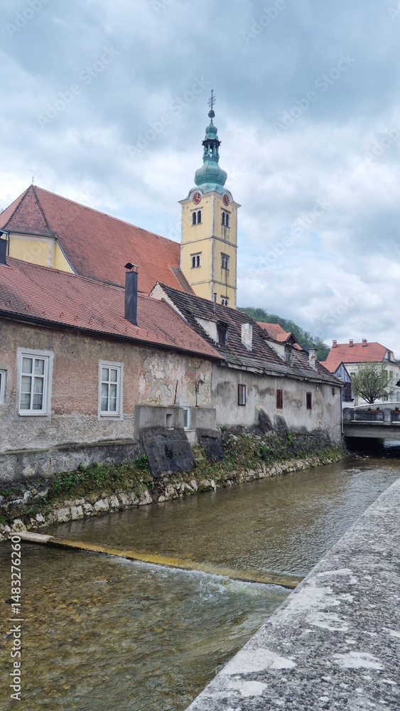 The steeple of St. Anastasia Church stands tall above the old, red-roofed buildings lining the river in Samobor under a cloudy sky