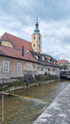 Obraz na płótnie The steeple of St. Anastasia Church stands tall above the old, red-roofed buildings lining the river in Samobor under a cloudy sky w salonie