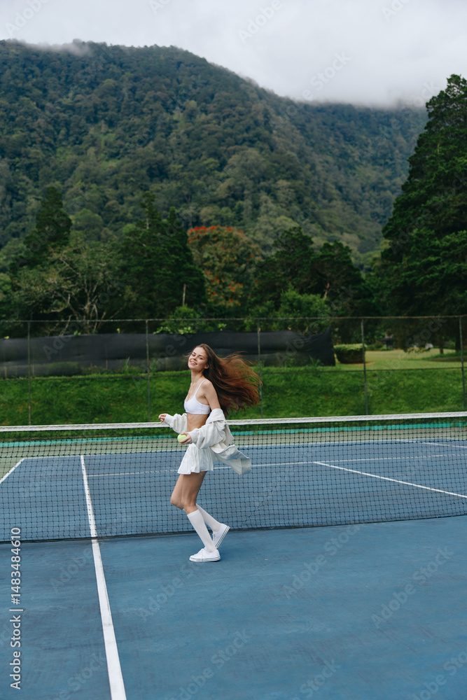 Obraz premium Beautiful Asian woman playing tennis on a sunny day, radiating joy and energy, surrounded by lush green nature and an inviting blue court