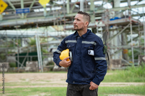 Surveyor or refinery worker in safety gear holding hardhat inspecting at oil petroleum factory. Engineer standing at construction site outdoor. Background of pipeline production