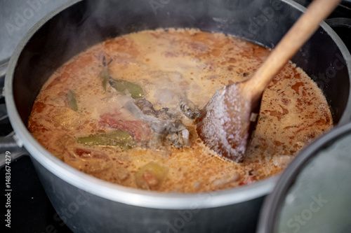 Top-down view of a pot filled with hearty soup, such as bograch or a creamy cheese-based soup. A wooden spoon rests over the pot, filled with steaming broth.