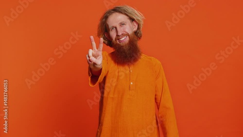 Happy Caucasian man showing victory sign, hoping for success and win, doing peace gesture, smiling with kind optimistic expression, celebrate. Redhead young guy isolated on studio orange background