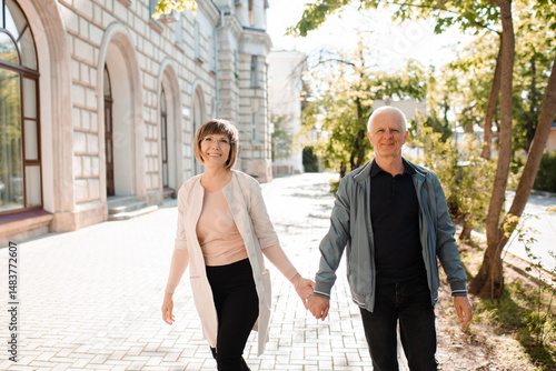 Senior happy couple of stylish man and woman 60-65 year old walking together on streets holding hands at city outdoors. Wellness lifestyle. Happiness.
