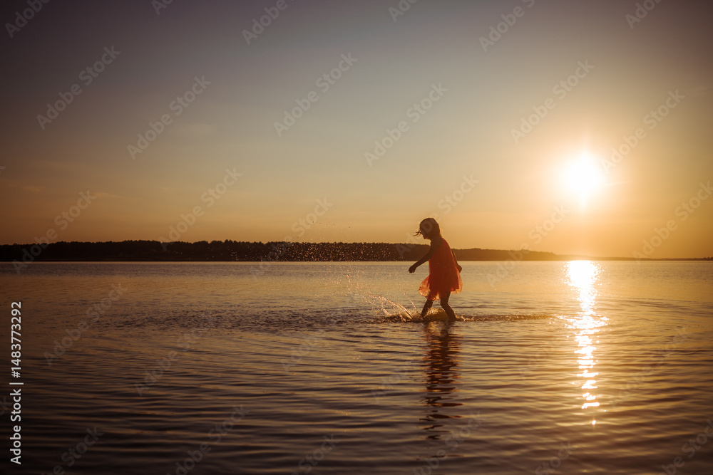 Fototapeta premium Silhouette of a girl running on water against the background of an evening sunset. Water splashes in all directions. Carefree baby fun. Happy healthy childhood.