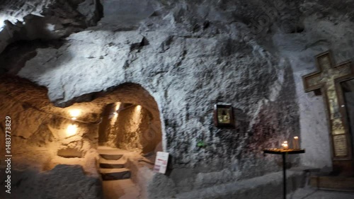 Interior of Vardzia, a 12th-century cave monastery in Georgia, showing a large wooden cross, candles, religious icon, and a staircase leading deeper into the rock-cut complex during a daytime visit