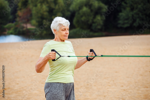 Old woman wear sportswear doing exercises outdoor on the beach in the morning using resistance rubber bands. Healthy lifestyle.