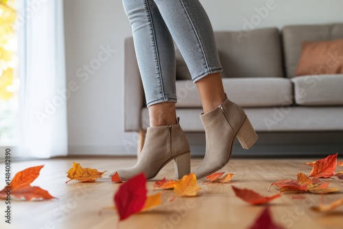 Woman wearing stylish ankle boots steps amid colorful autumn leaves on a wooden floor in a cozy room.